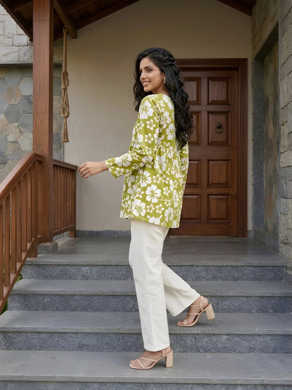 A side-profile, full-length shot of a woman in a green floral tunic and white pants, looking over her shoulder while walking up stone steps toward a wooden door.