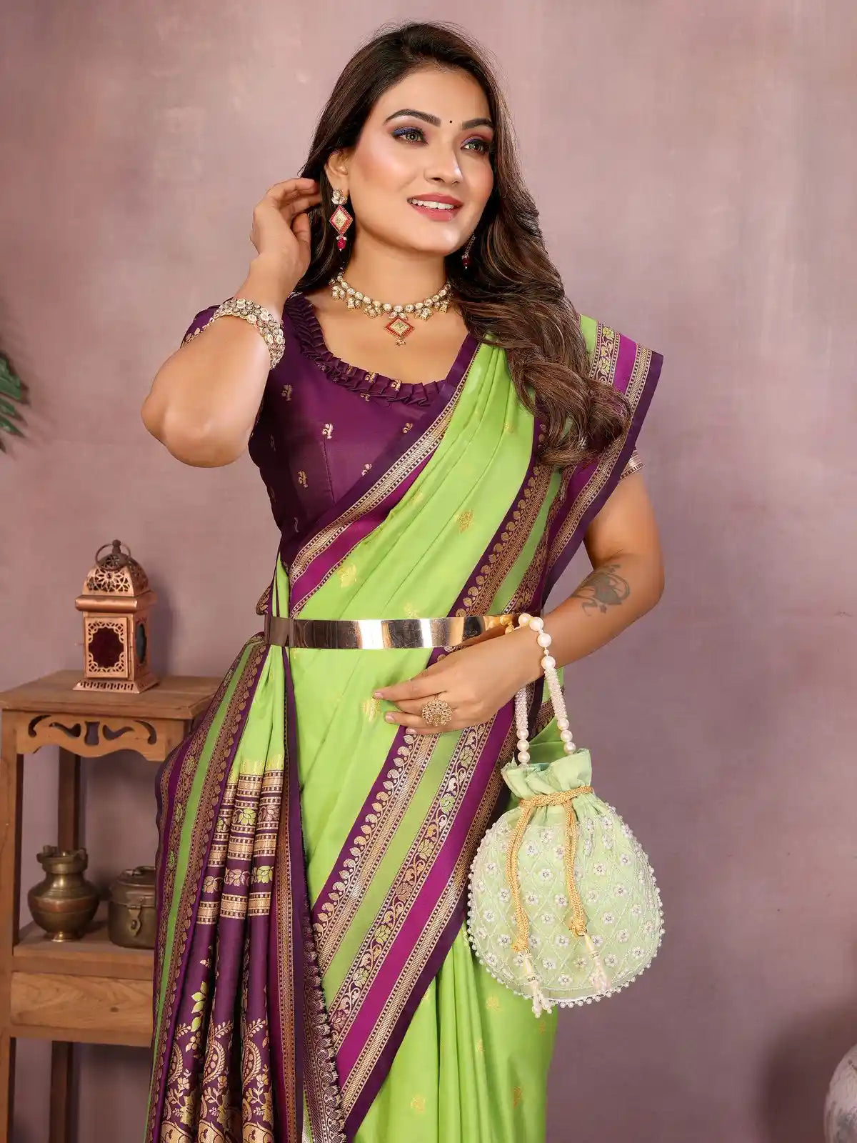 Woman wearing a green and purple saree with jewelry and a handbag against a neutral background