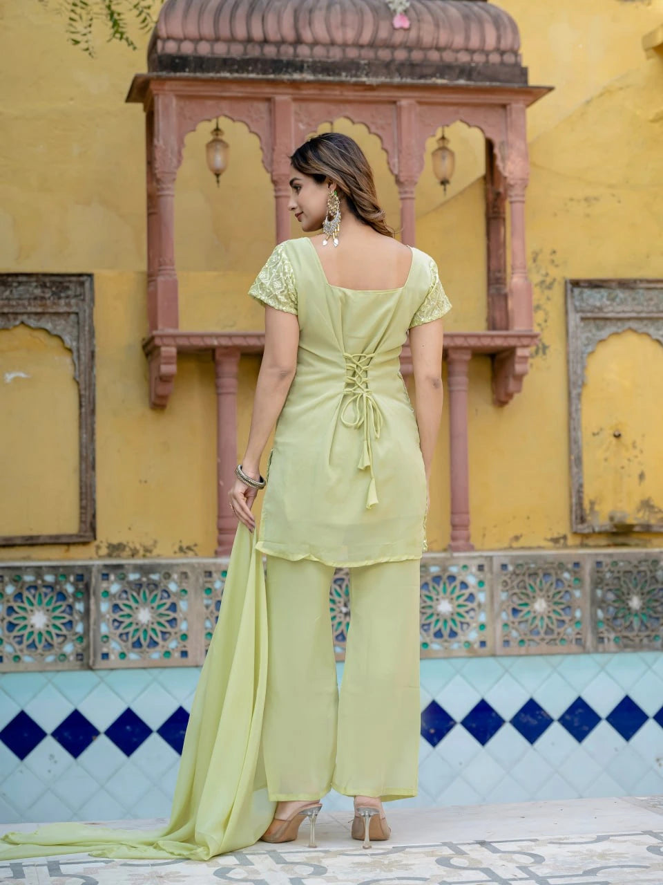 Woman in a light green traditional outfit standing in front of a decorative wall with arches and tiles.