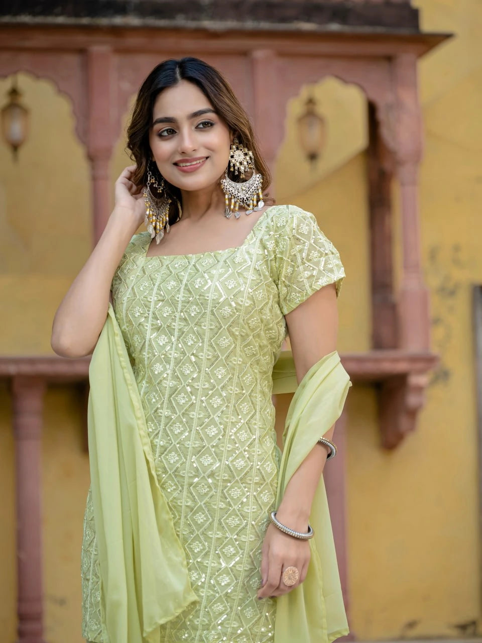 Woman in a light green traditional outfit with jewelry against a yellow architectural background