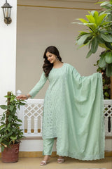 Woman in a light green traditional outfit standing outdoors with plants around.