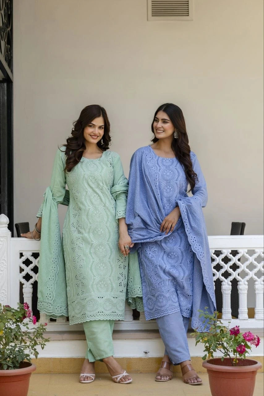 Two women in traditional outfits standing on a balcony with plants and a white railing.