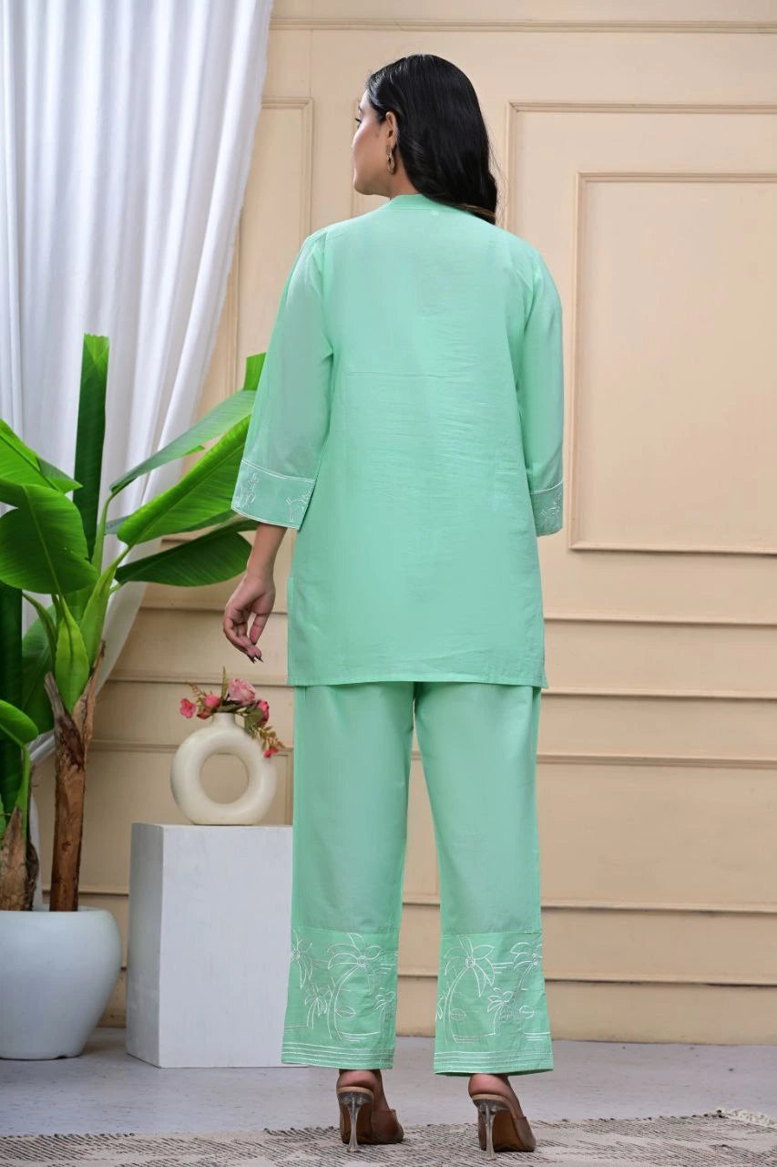 Woman wearing a light green traditional outfit with embroidery, standing in front of a beige wall.