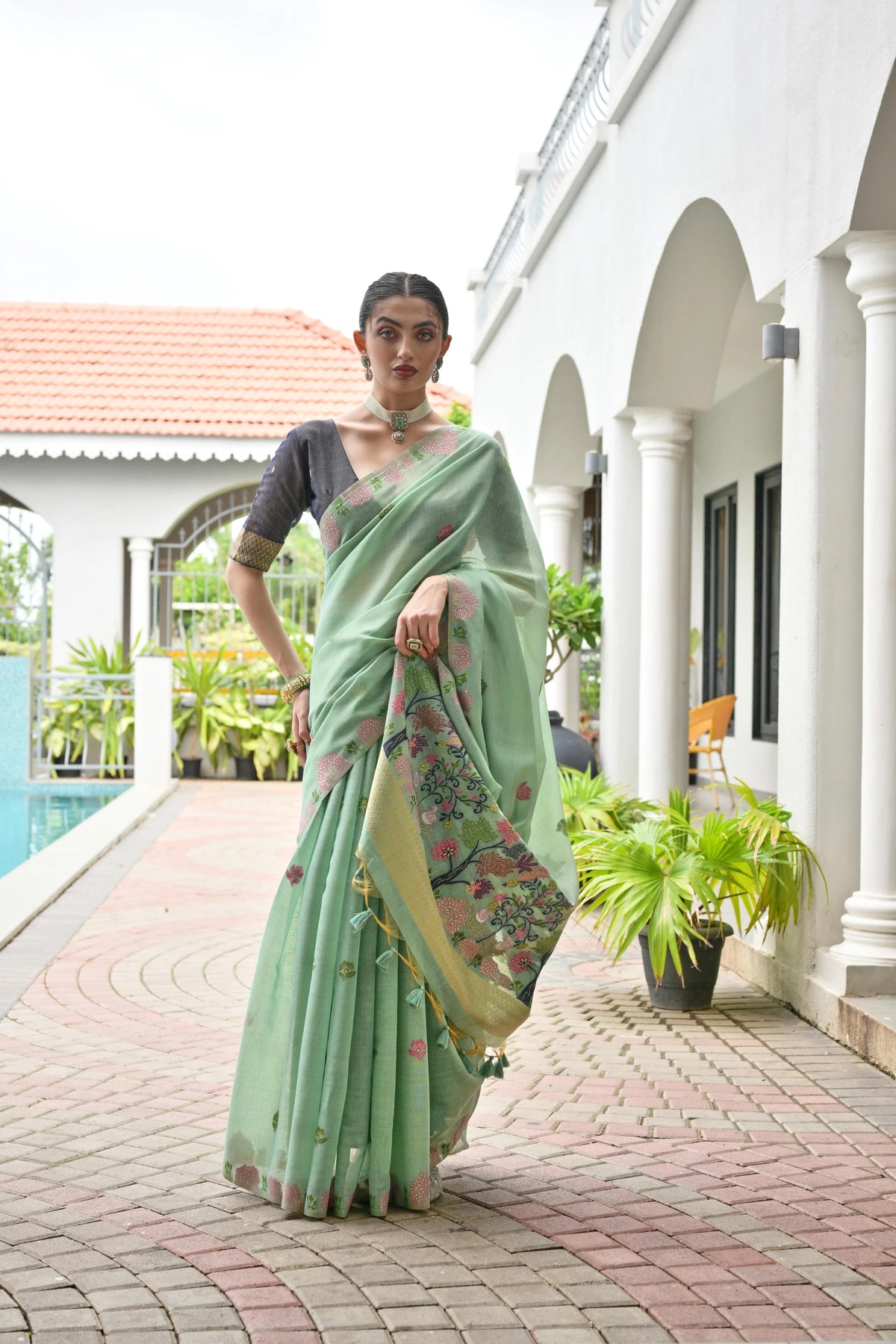 Woman in a light green saree with a floral pattern standing in front of a poolside building.
