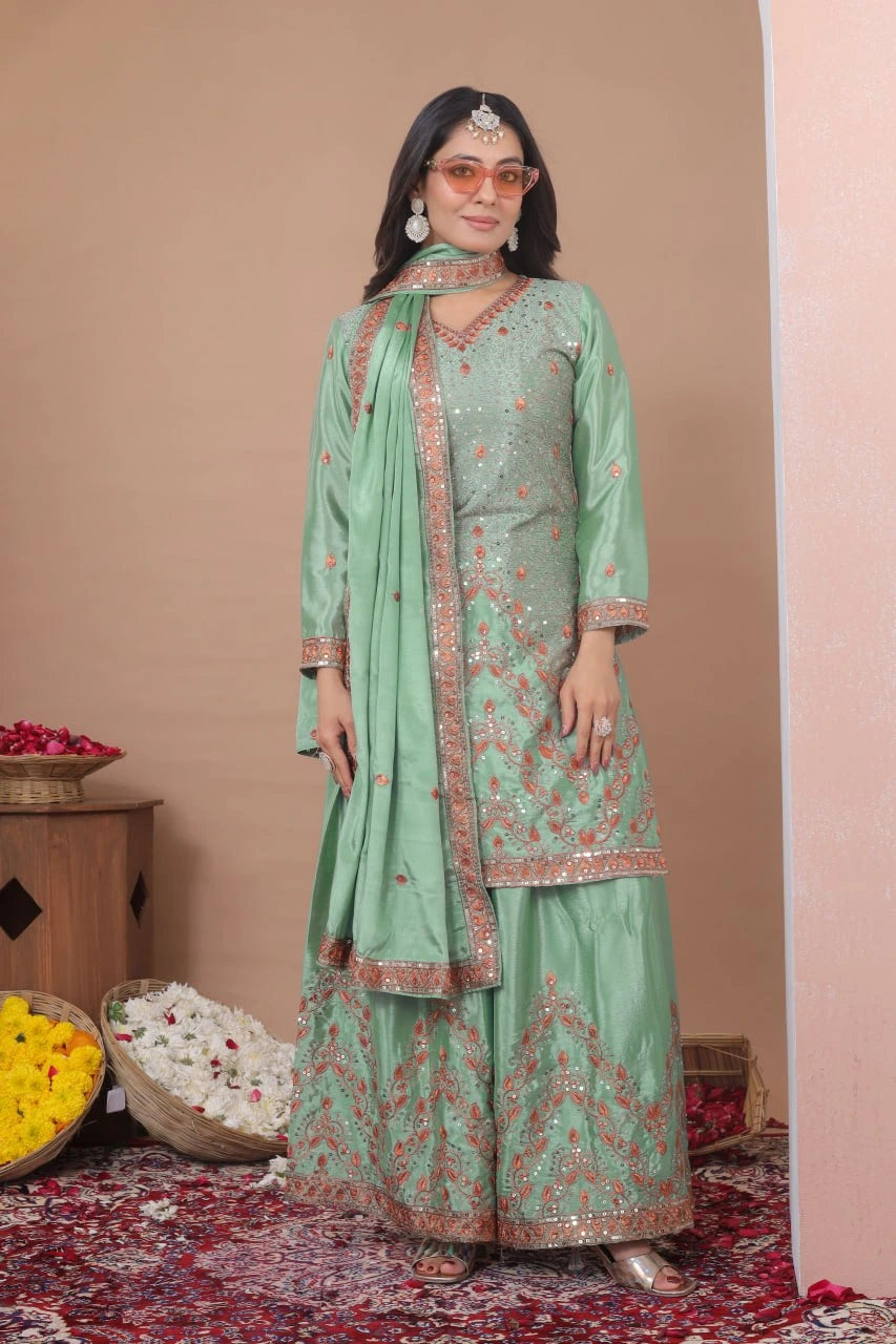 A woman is shown from a back-profile view, looking over her shoulder with a smile. She is wearing a light green ethnic suit featuring wide-legged pants and a matching dupatta, all adorned with intricate orange and silver embroidery along the borders. The setting includes baskets of rose petals and yellow flowers on a patterned rug against a neutral background.