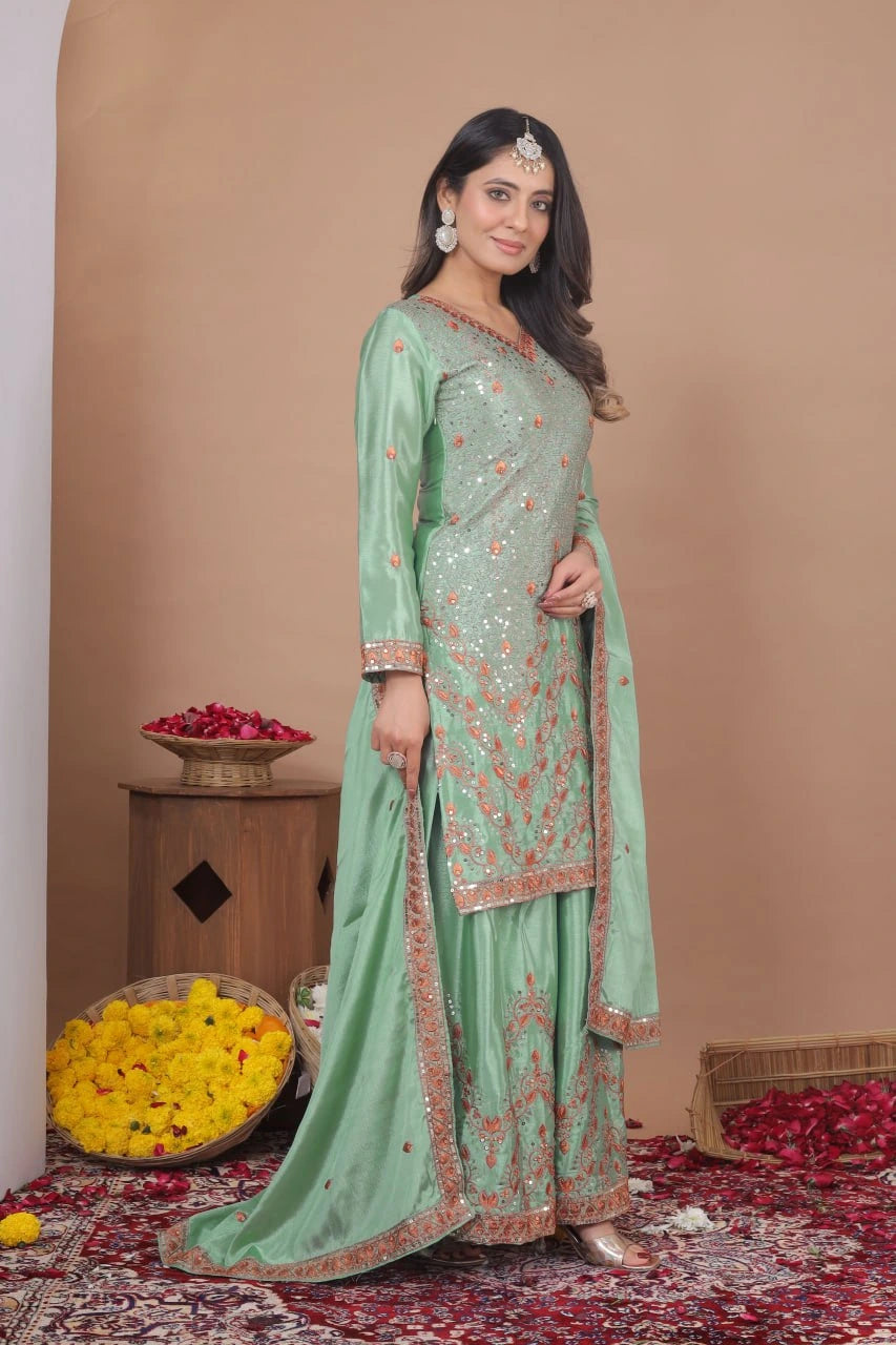 Woman in a light green traditional outfit with intricate patterns, standing against a beige wall with decorative elements.