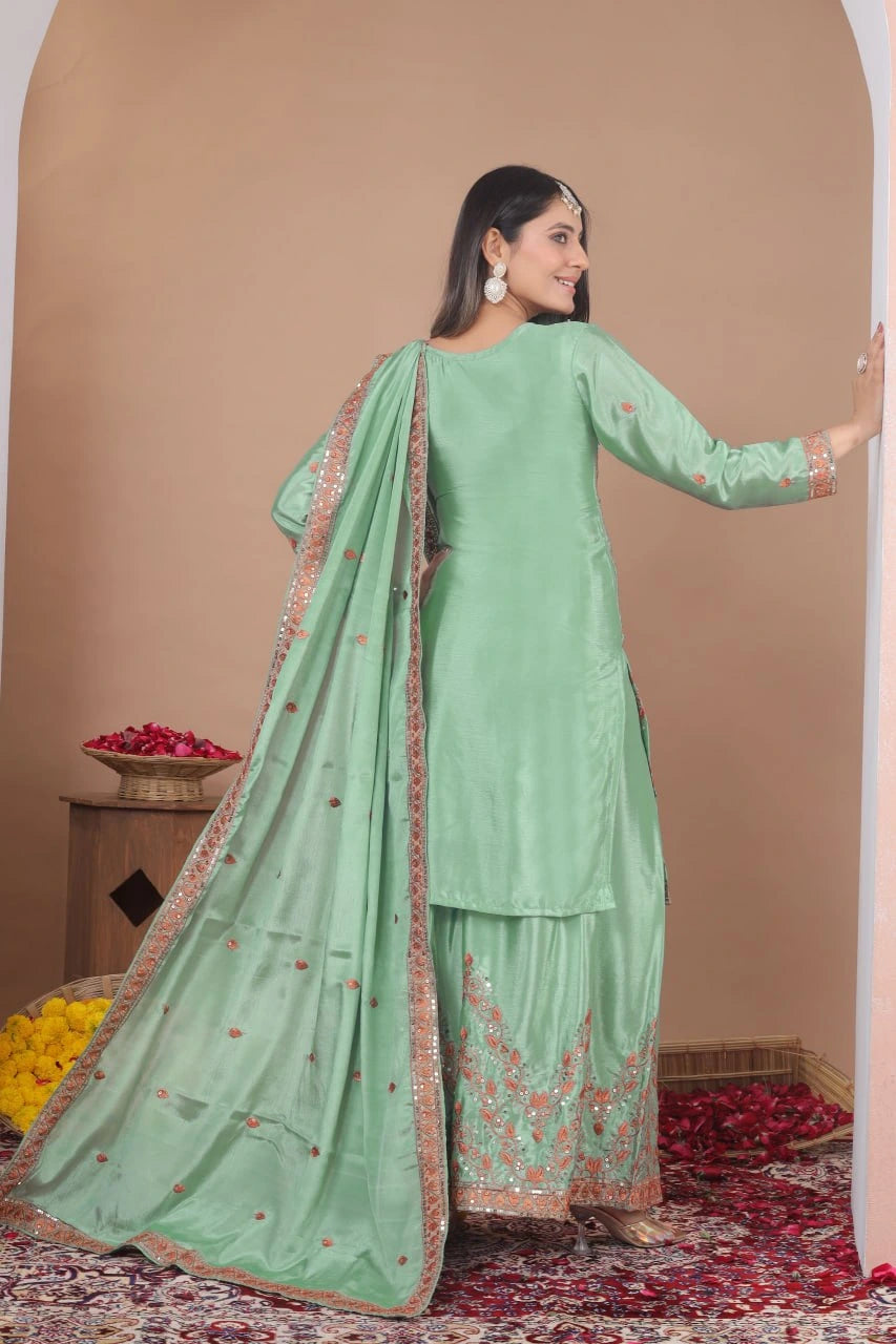 A front-facing view of a woman wearing a light green kurta and sharara set with heavy orange and silver floral embroidery. She is accessorized with orange-tinted sunglasses, large silver earrings, and a traditional headpiece (maang tikka). She stands in a decorated indoor space with baskets of colorful flowers at her feet.