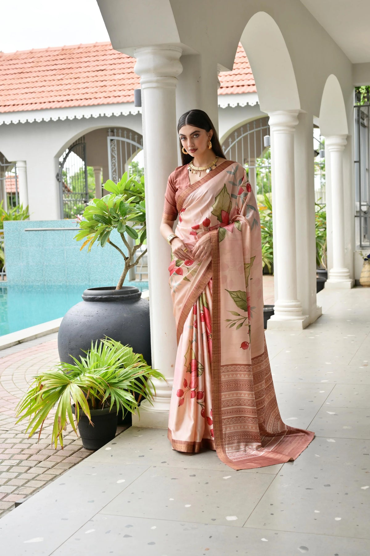 Woman in a floral saree standing by a poolside with white columns and plants.