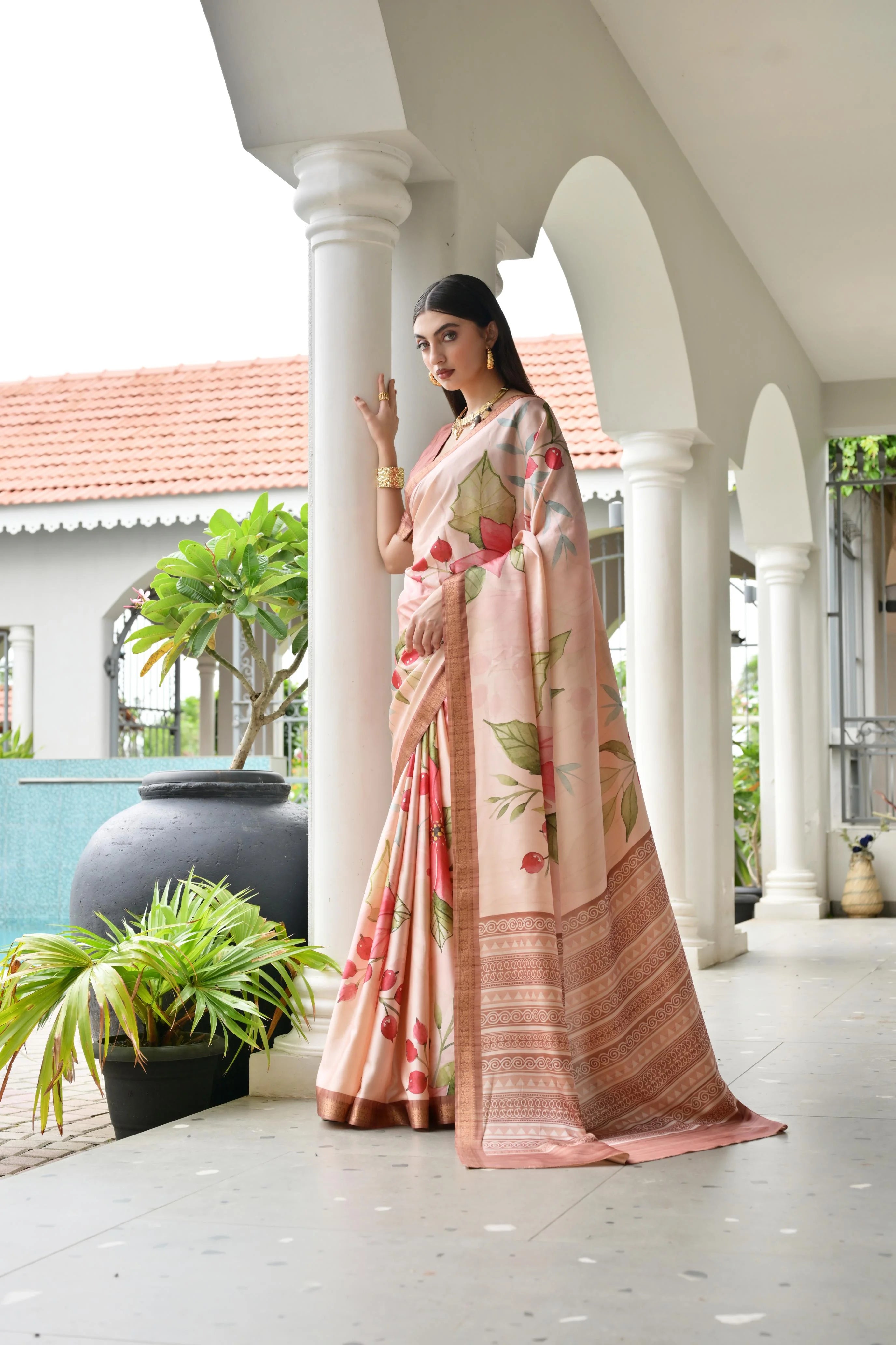 Woman in a floral saree standing in a decorative outdoor setting with columns and plants.