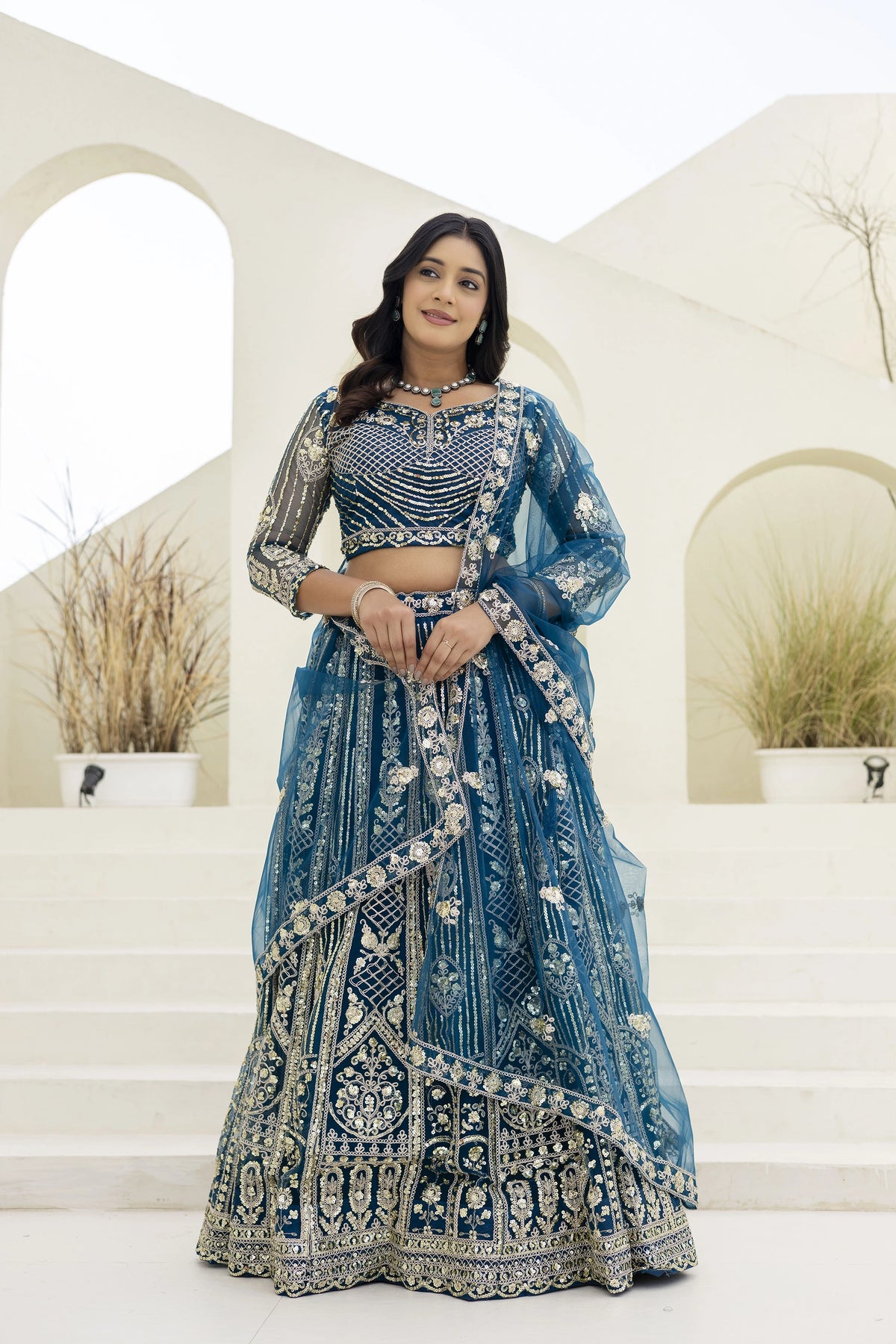 Woman in a blue embroidered traditional outfit standing in front of decorative arches.