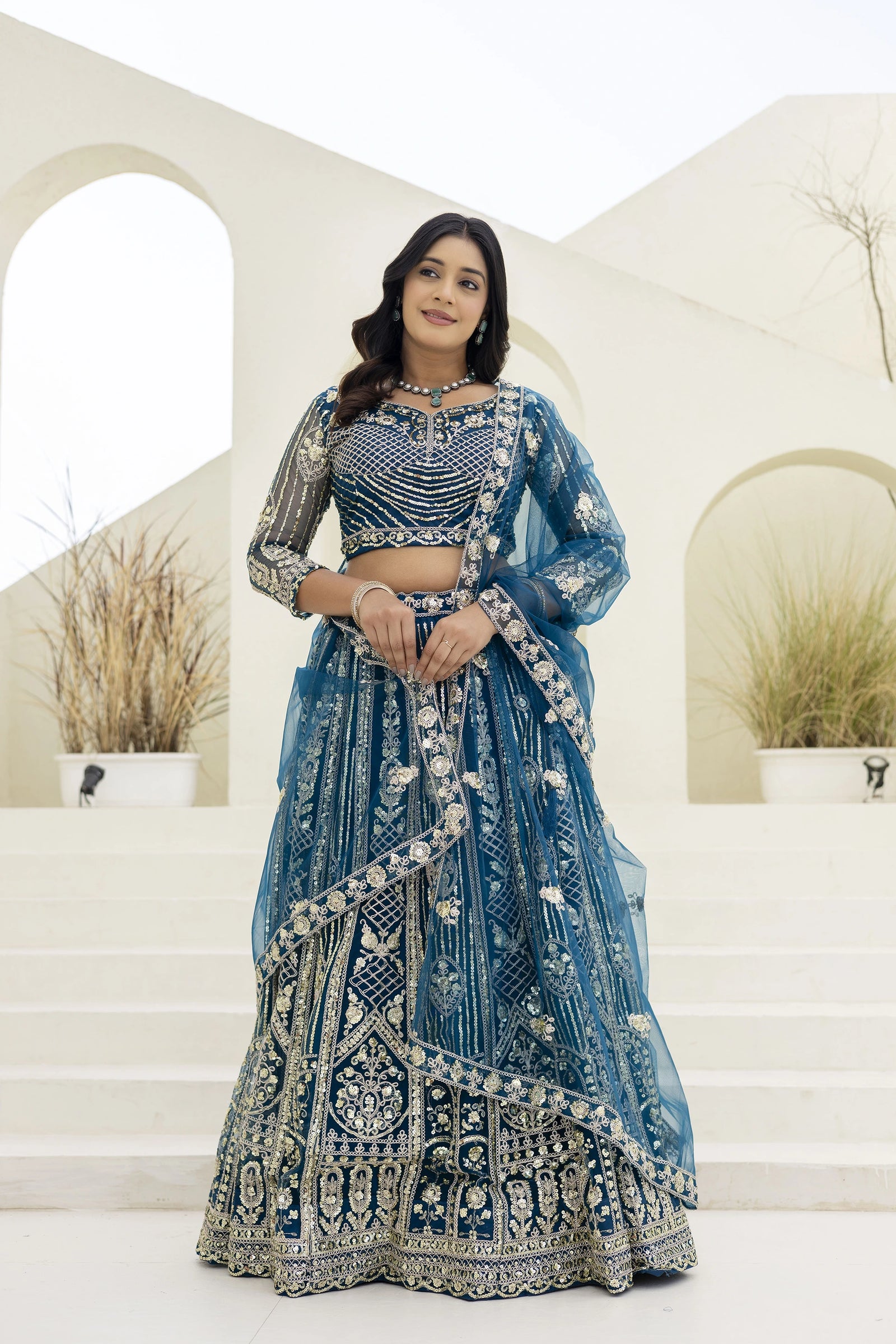 Woman in a blue embroidered traditional outfit standing in front of decorative arches.