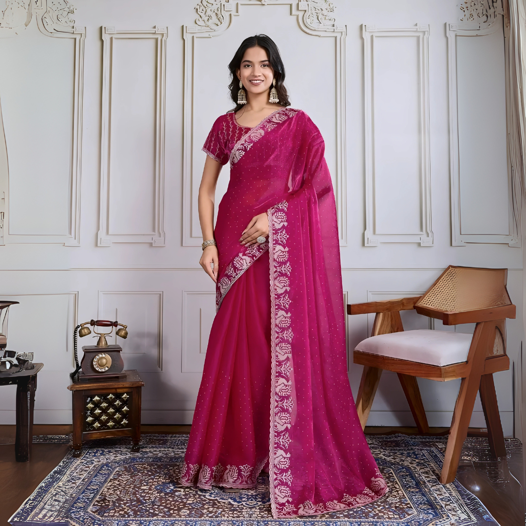 Woman wearing a magenta georgette saree adorned with subtle sequins and an ornate silver embroidered floral border, paired with a matching embroidered blouse. She stands elegantly in a classic white paneled room, accessorized with statement jhumka earrings and bangles, next to a vintage rotary telephone on a carved wooden side table and a modern wooden armchair set atop a decorative patterned rug for a graceful, timeless look.