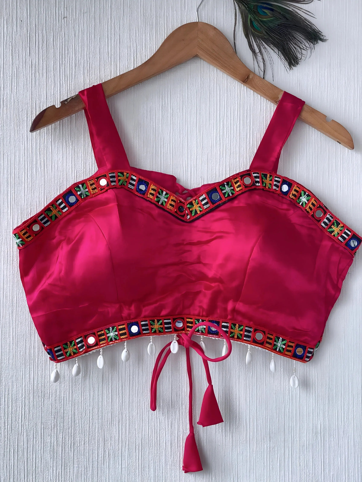  Fuchsia pink ethnic blouse featuring vibrant mirror work and a multicolored embroidered geometric border in green, blue, and orange, adorned with pearl drop tassels and a tie-up waist detail. Displayed on a wooden hanger against a textured off-white wall with peacock feathers, creating a festive and traditional look.