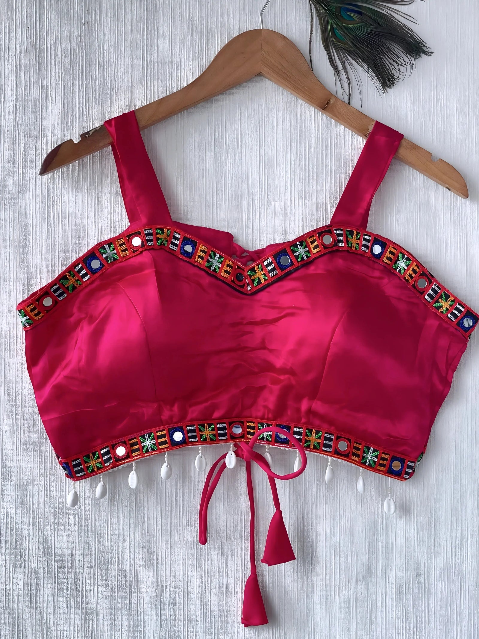  Fuchsia pink ethnic blouse featuring vibrant mirror work and a multicolored embroidered geometric border in green, blue, and orange, adorned with pearl drop tassels and a tie-up waist detail. Displayed on a wooden hanger against a textured off-white wall with peacock feathers, creating a festive and traditional look.