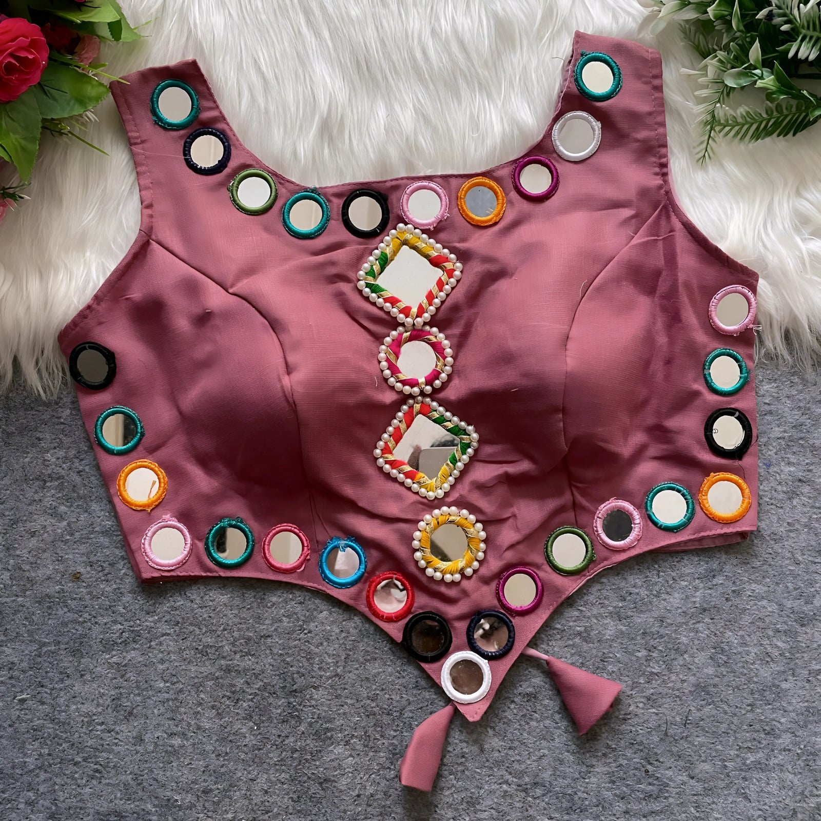 Mauve ethnic blouse featuring vibrant mirror work with multicolored embroidered borders and decorative square and round mirrors arranged vertically at the center, accented with elegant pearl detailing and tie-up tassels at the bottom. Displayed flat on a grey surface with a white faux fur background and green leafy accents, highlighting its festive and traditional design.