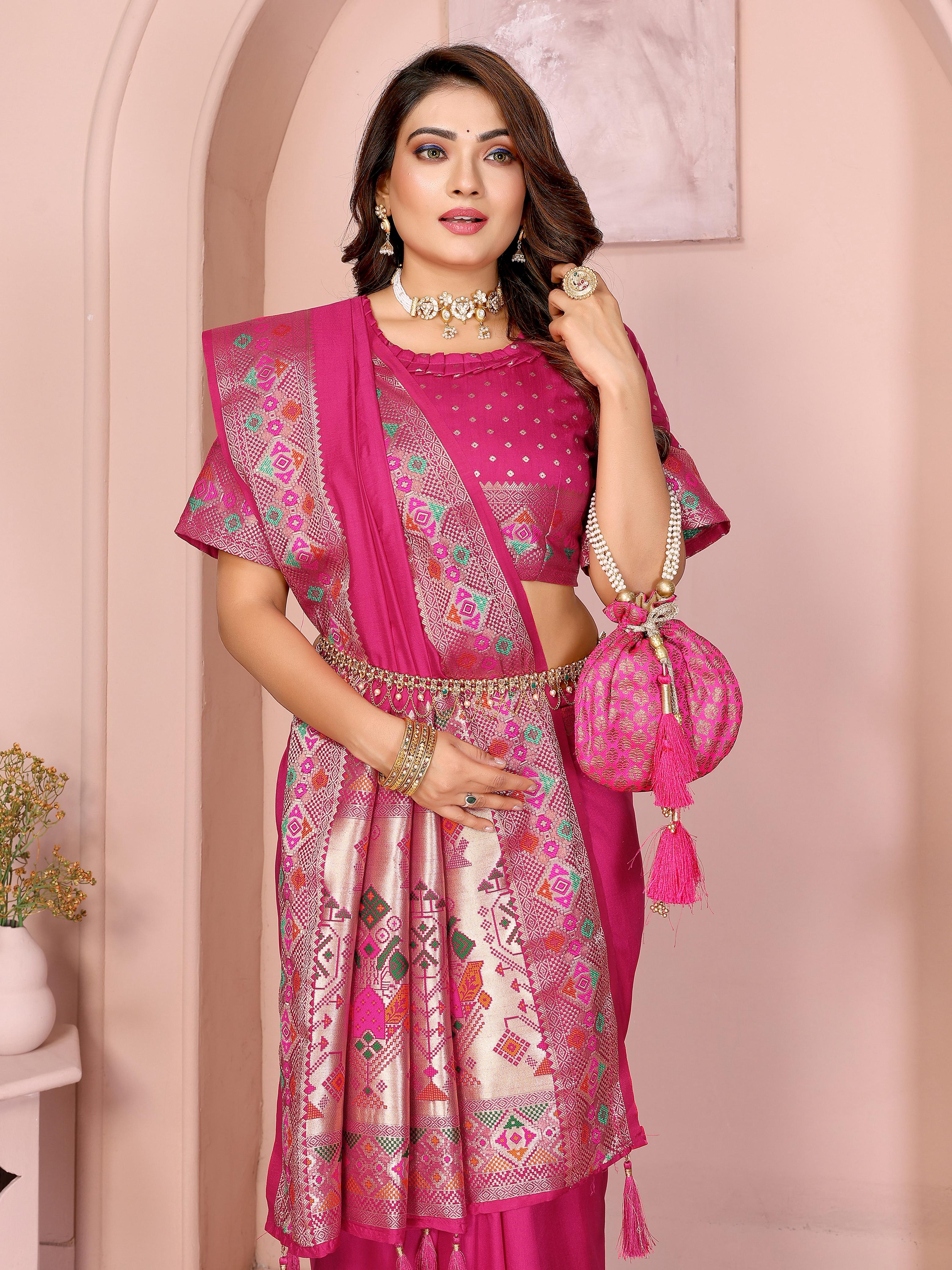 Woman wearing a magenta silk saree featuring a contrasting ethnic border with intricate geometric and floral patterns in gold and multi-color detailing, and matching tassels on the pallu. She pairs it with a matching blouse showcasing rich embroidery. She accessorizes with traditional choker, necklace, and bangles, standing elegantly in a pastel arched studio with potted plants and minimalist decor, presenting a refined and graceful ethnic look.