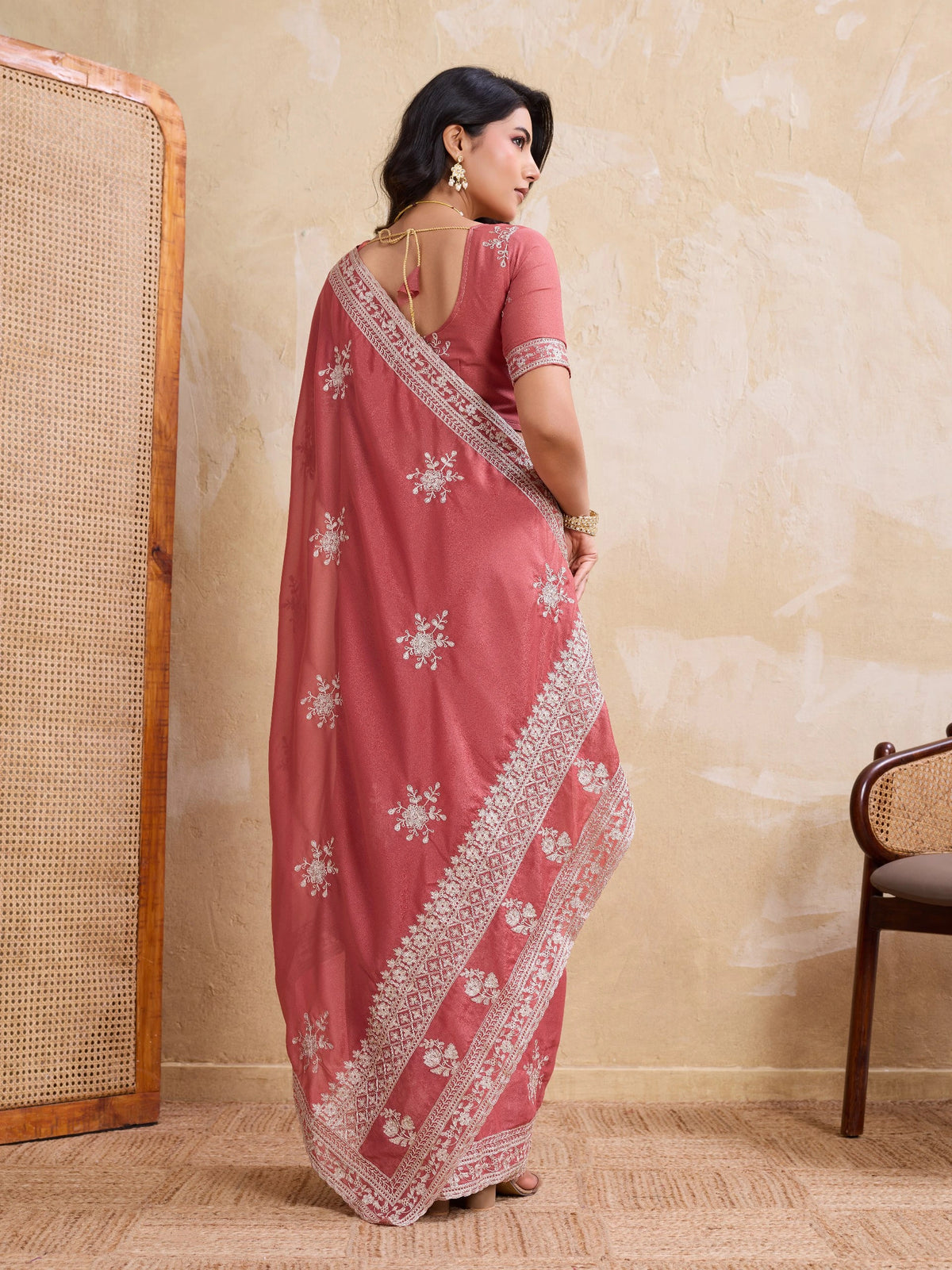 Woman wearing a pink saree with white patterns against a beige wall.