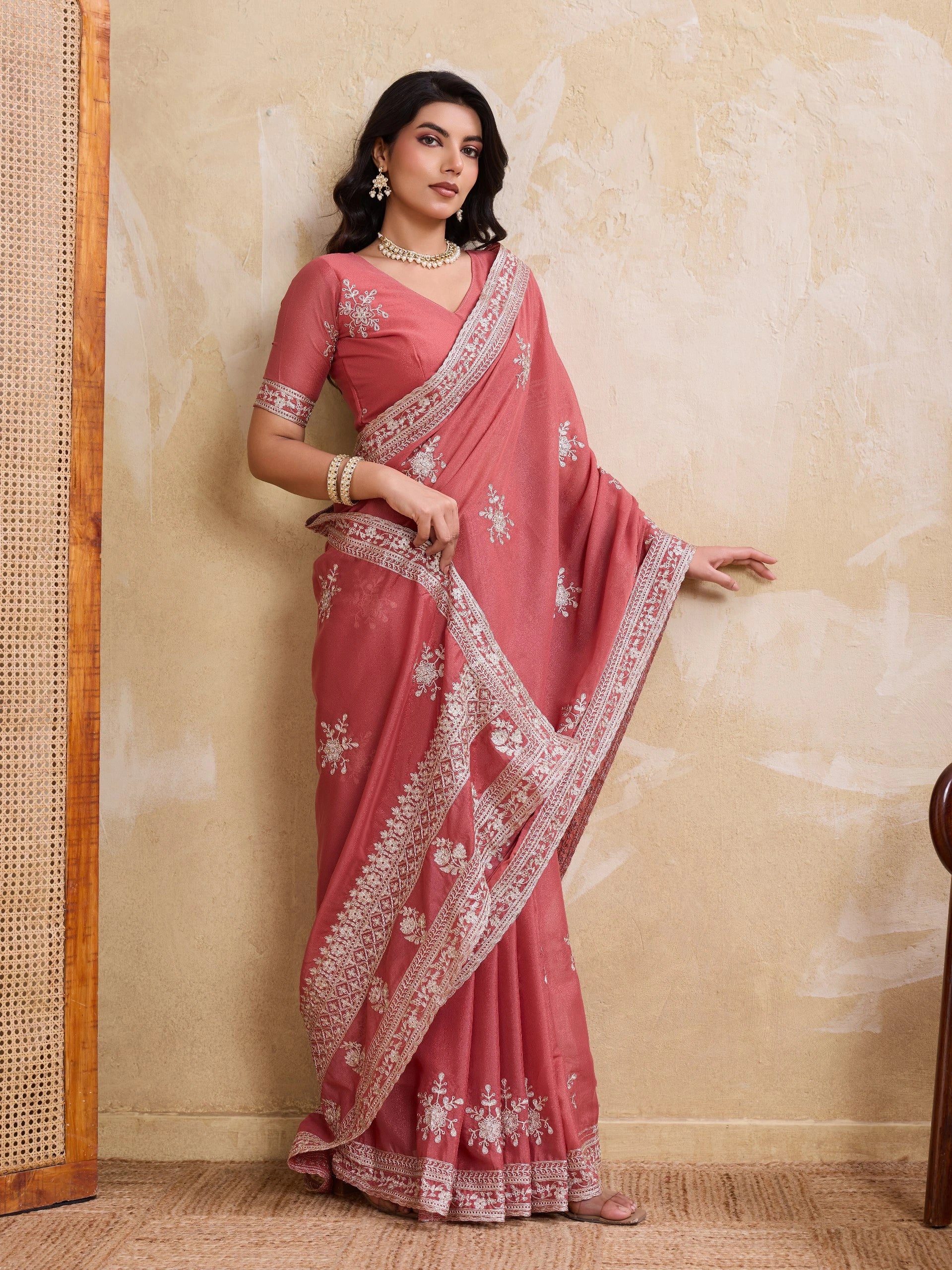 Woman wearing a pink saree with white embroidery against a beige wall.