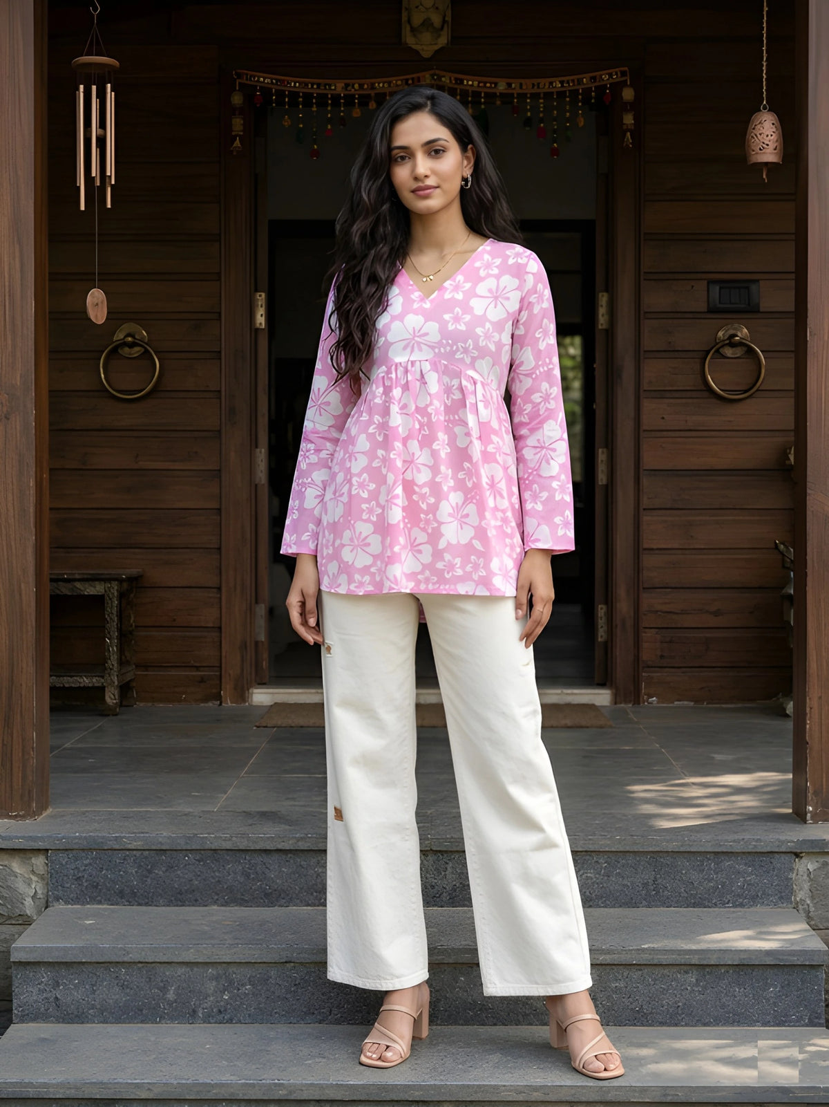 A full-length shot of a woman in a pink floral-print peplum tunic and white wide-leg pants standing in front of a wooden house entrance.