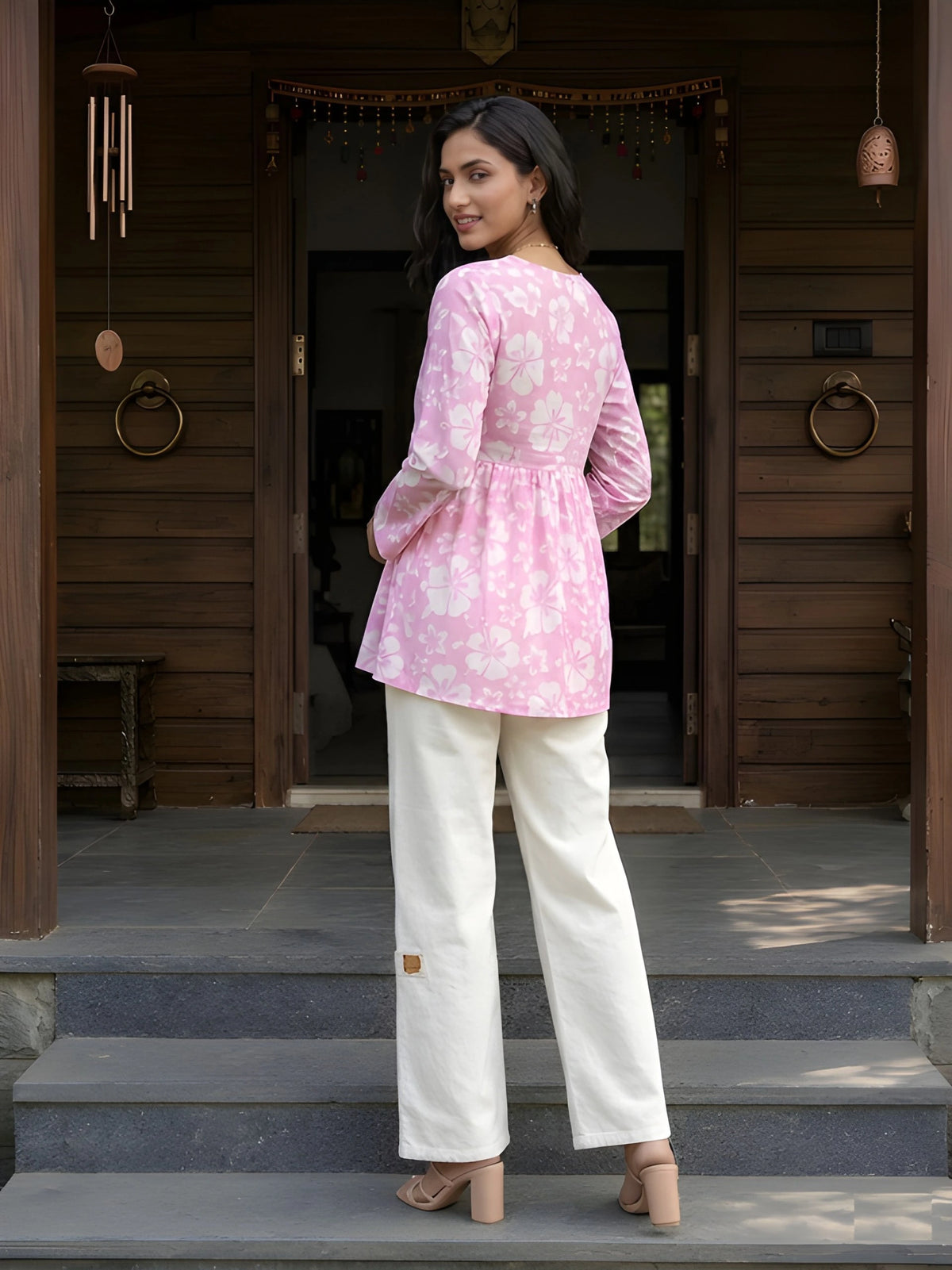 A full-length shot from behind of a woman looking back over her shoulder, showing the design of her pink floral tunic and white pants.