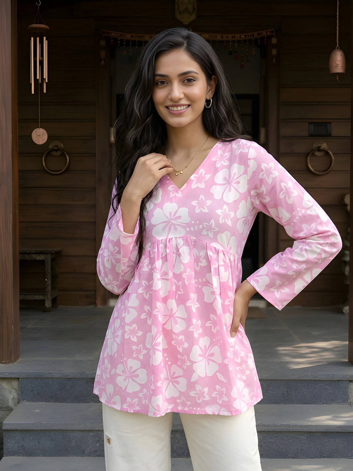 A medium shot of a smiling woman with long dark hair, wearing a pink floral peplum top and touching a gold necklace.