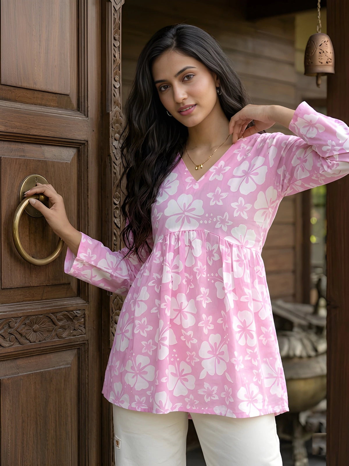A medium shot of a woman with long wavy hair posing with her hand on her head, wearing a pink floral tunic