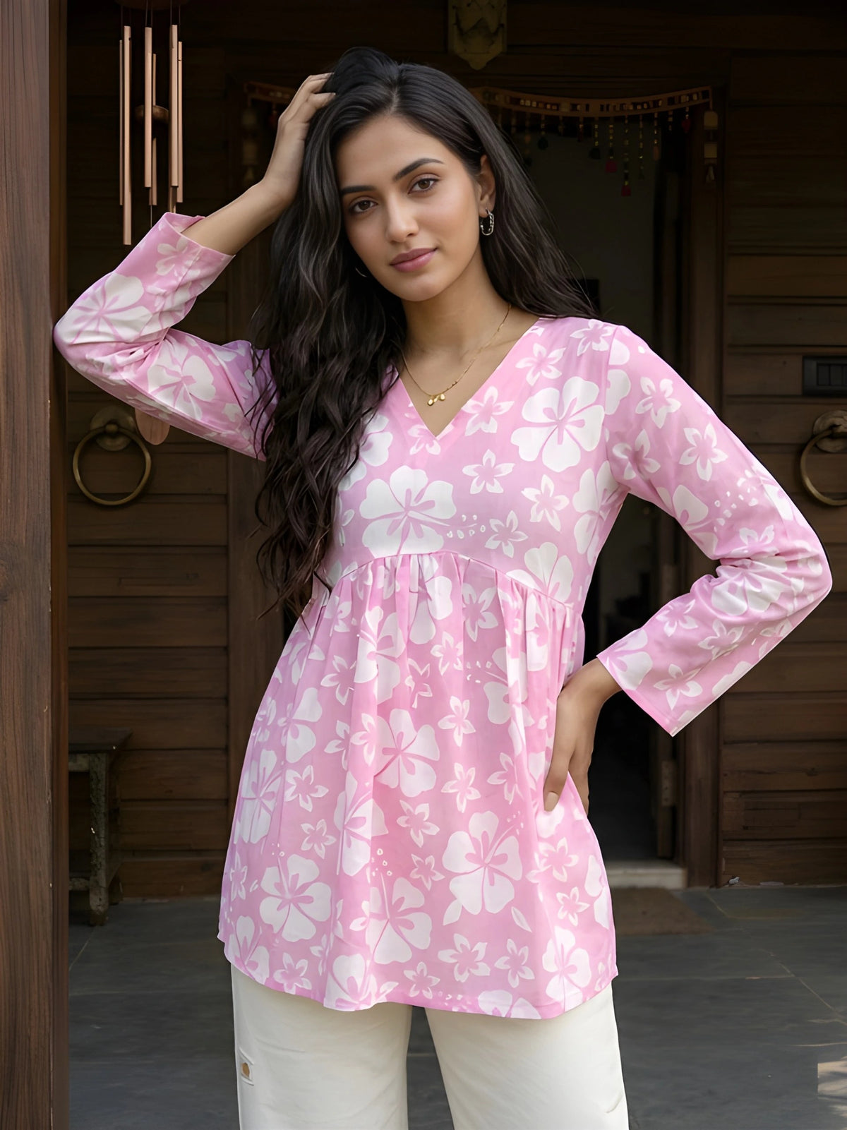 A medium shot of a woman leaning against a dark wooden door and holding a large brass ring handle while wearing a pink floral top.