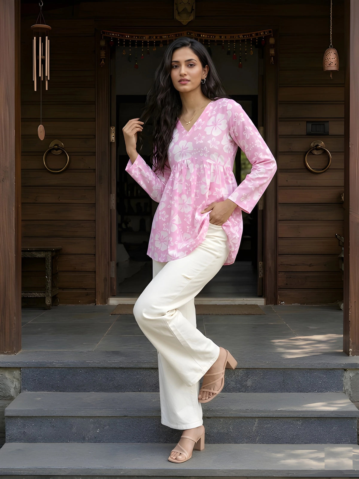 A full-length shot of a woman posing with one leg lifted and a hand in her pocket, dressed in a pink floral tunic and white trousers.