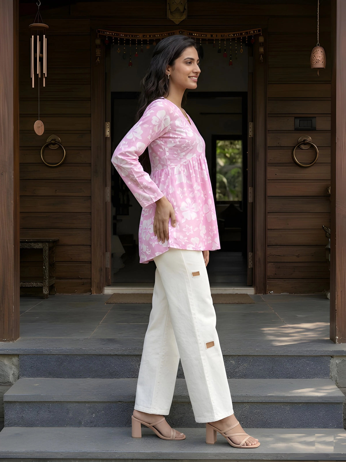 A full-length side-profile shot of a woman smiling and walking across stone steps in front of a wooden house entrance.