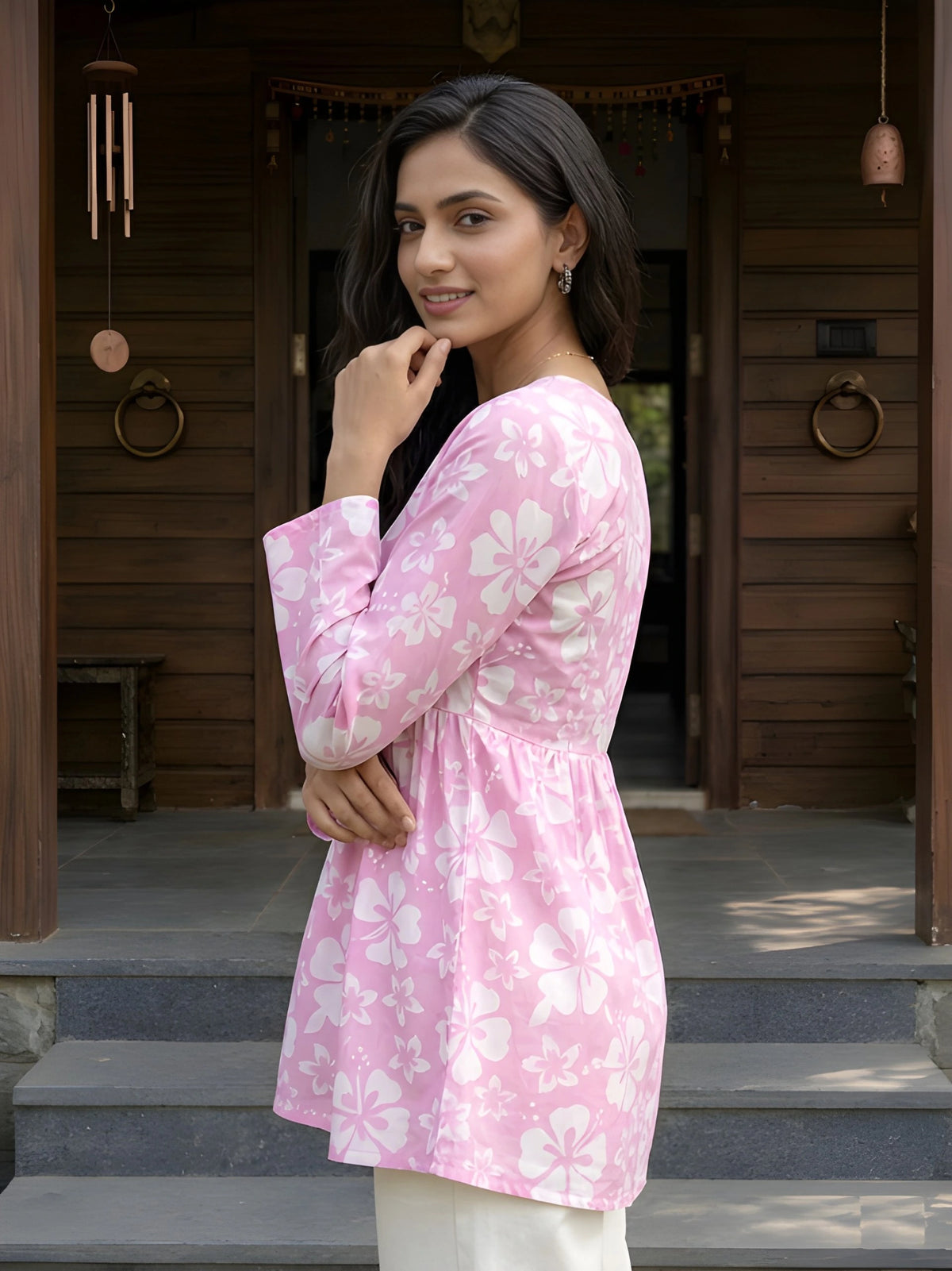 A side-profile medium shot of a woman wearing a pink floral tunic, looking toward the camera with her hand on her chin.