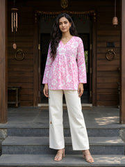 A full-length shot of a woman in a pink floral-print peplum tunic and white wide-leg pants standing in front of a wooden house entrance.