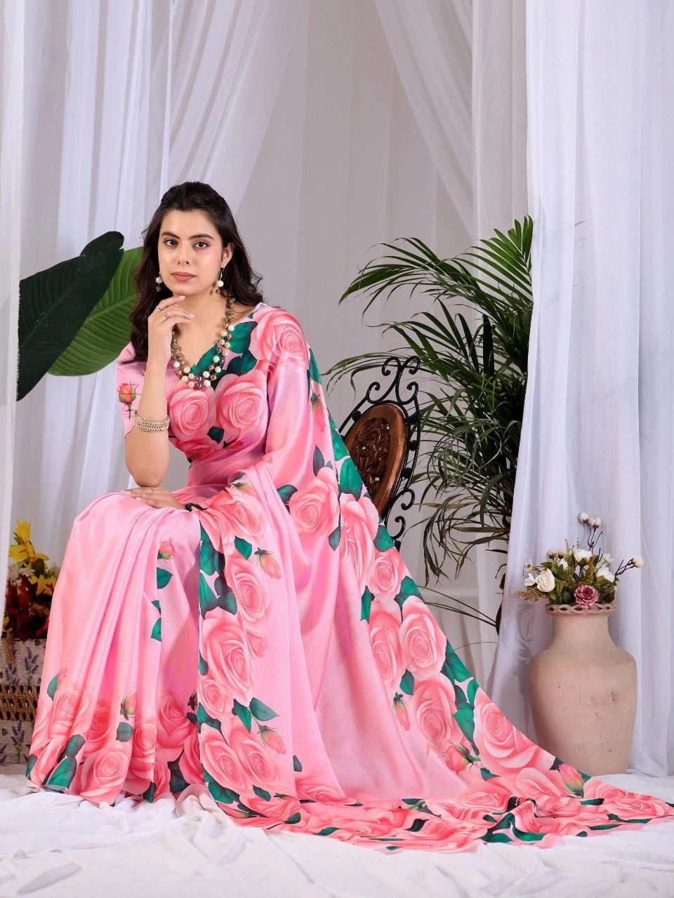 Woman in a pink floral saree sitting in a decorated room with plants and flowers.