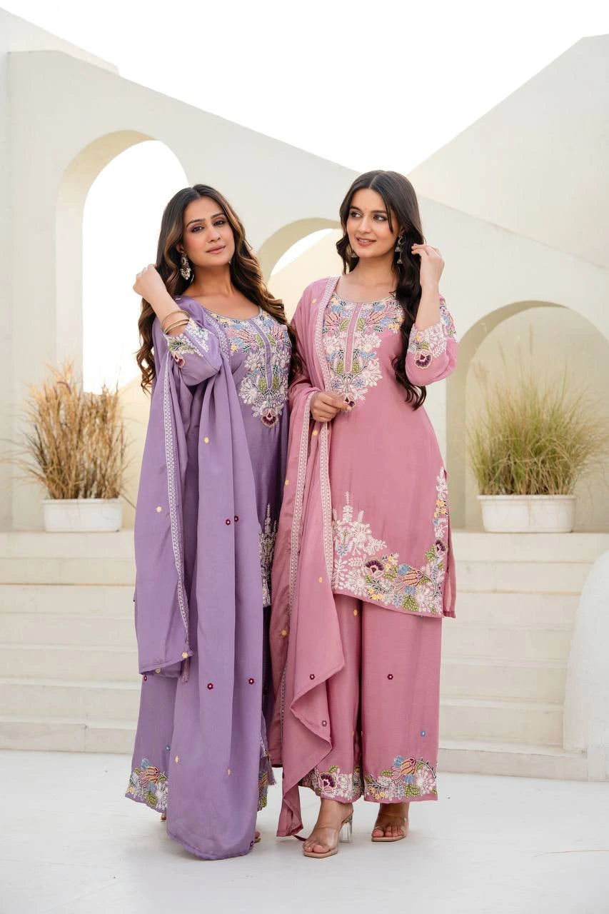 Two women in traditional outfits standing in a decorative setting with plants.