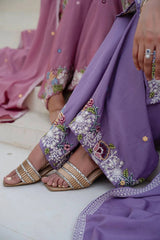 Person wearing a purple embroidered outfit with sandals on a light background