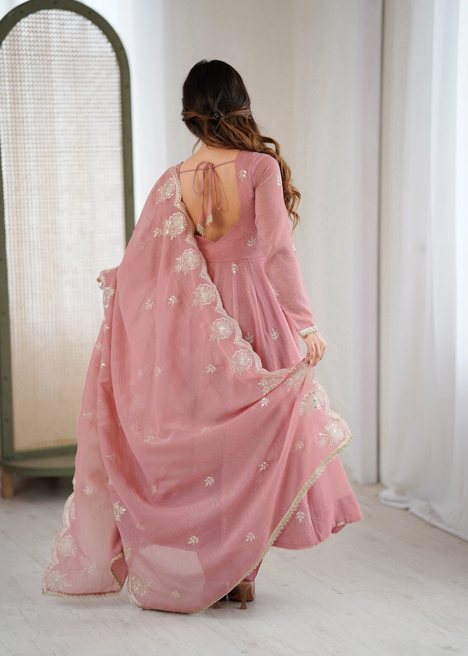 Woman wearing a pink saree with white floral embroidery in a minimal indoor setting.