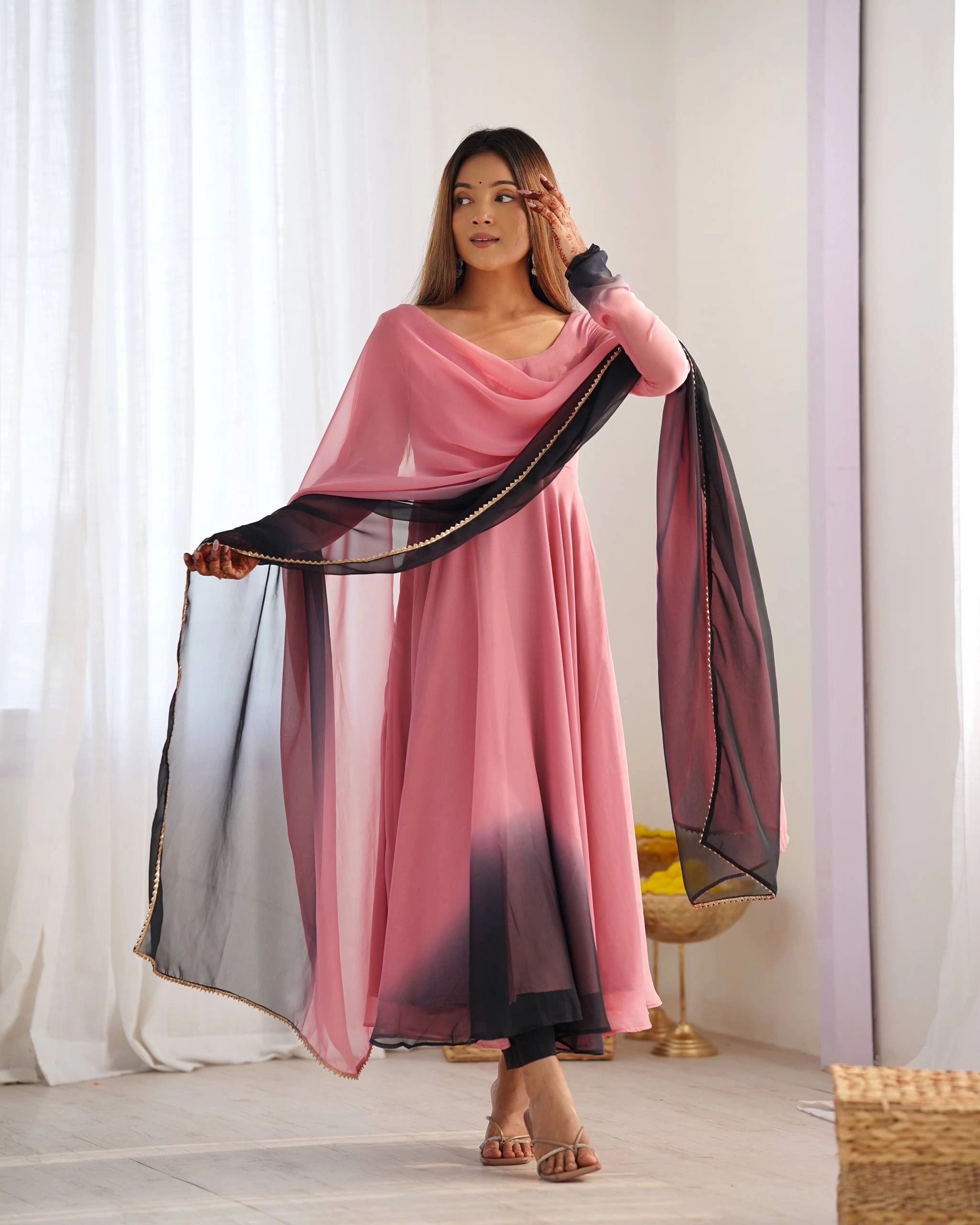 A woman in a pink and black ethnic suit stands with one hand near her forehead, looking to the side. She holds the edge of her dual-toned dupatta.