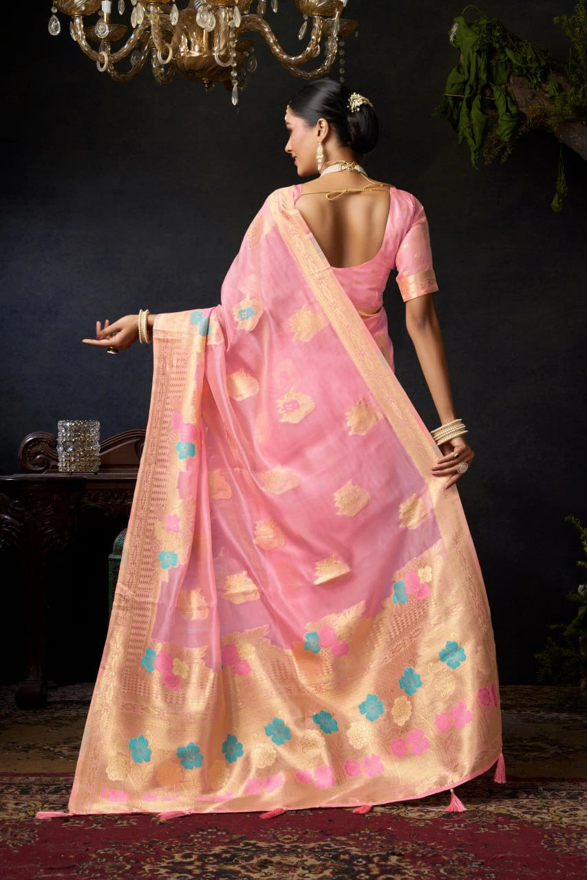 Woman in a pink saree with gold and blue patterns, standing against a dark background.