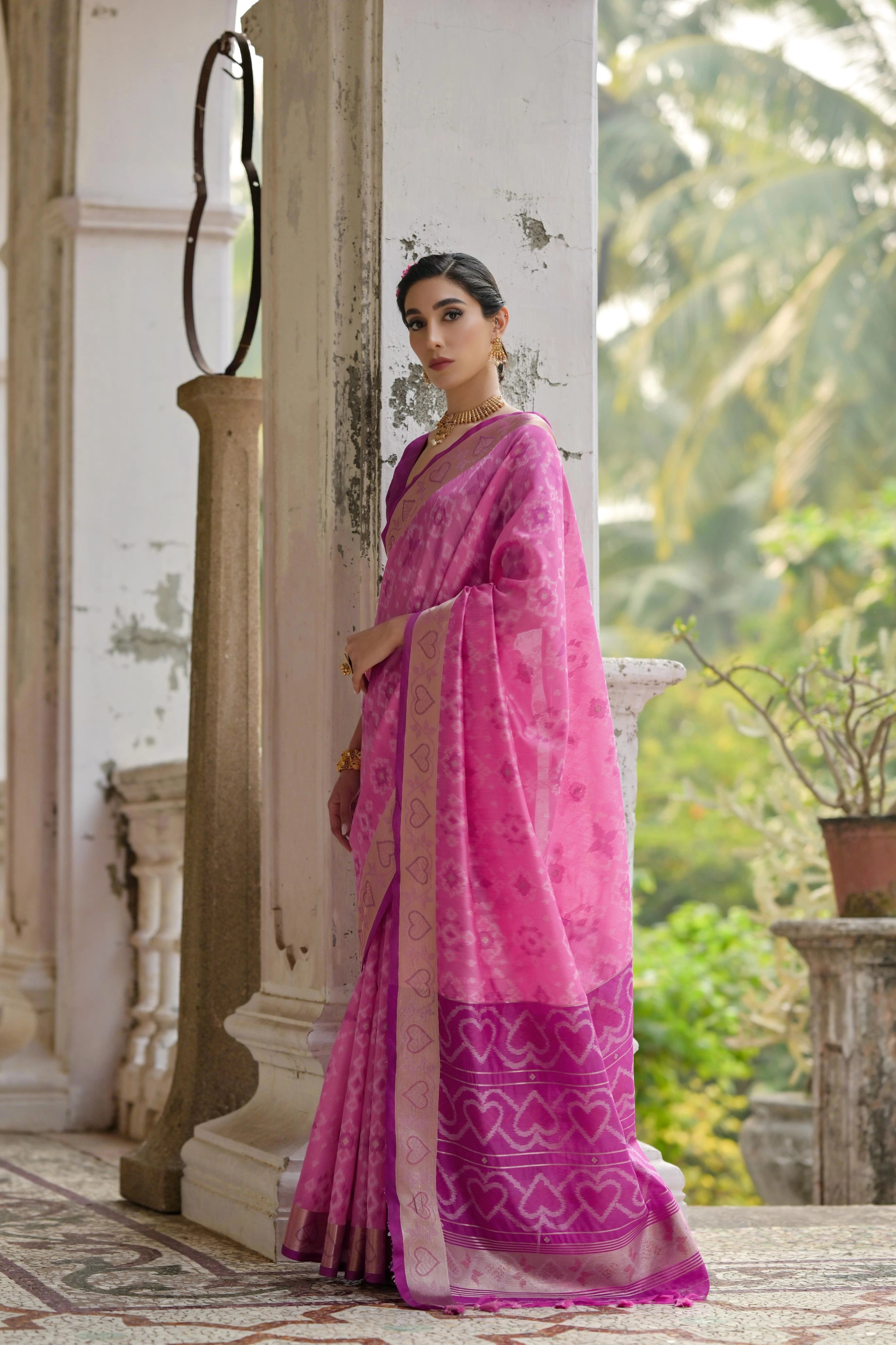 Woman in a pink saree standing in an outdoor setting with greenery.