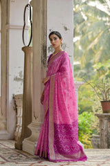 Woman in a pink saree standing in an outdoor setting with greenery.
