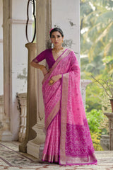 Woman in a pink saree standing in an outdoor setting with greenery.