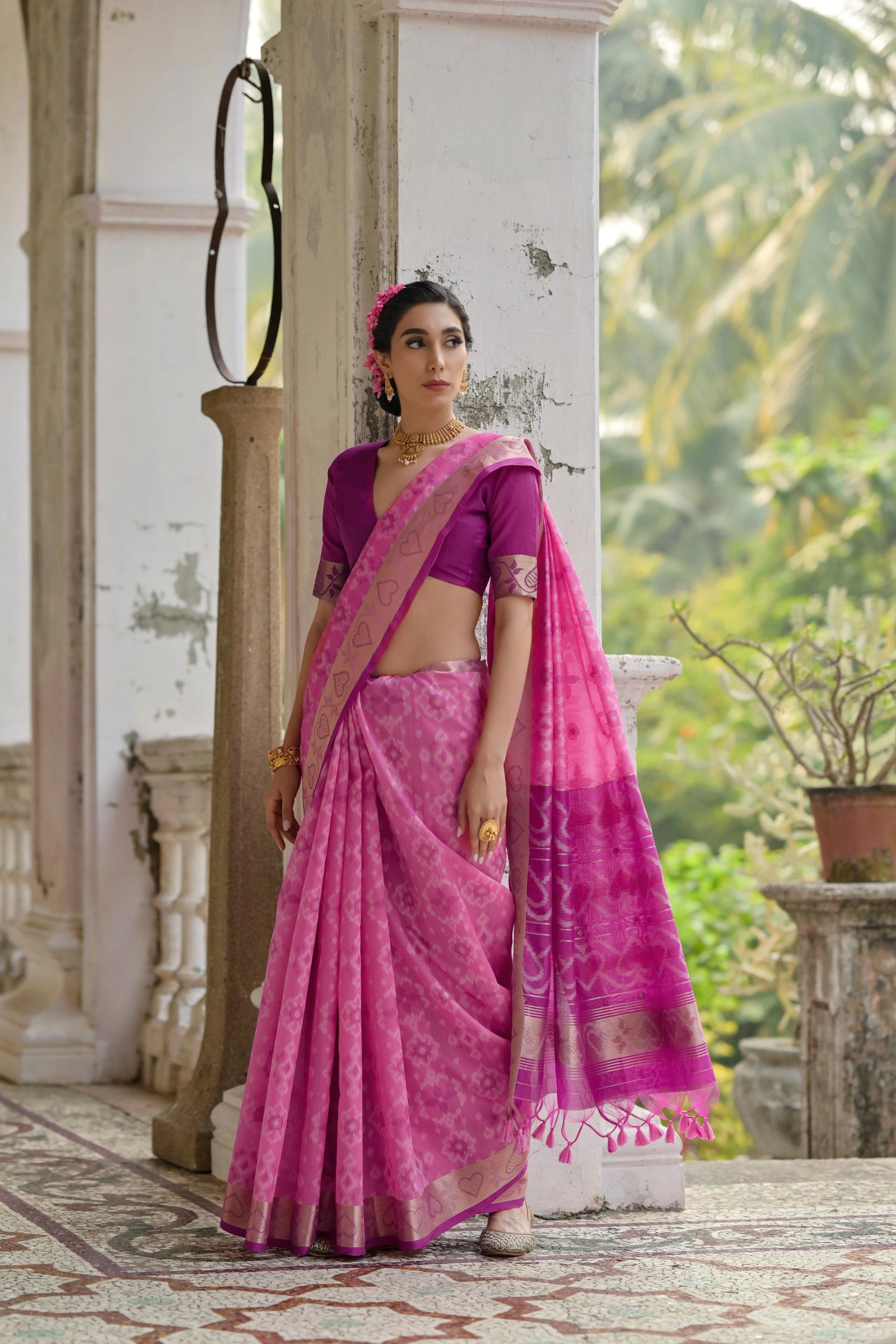 Woman in a pink saree standing in an outdoor setting with greenery.