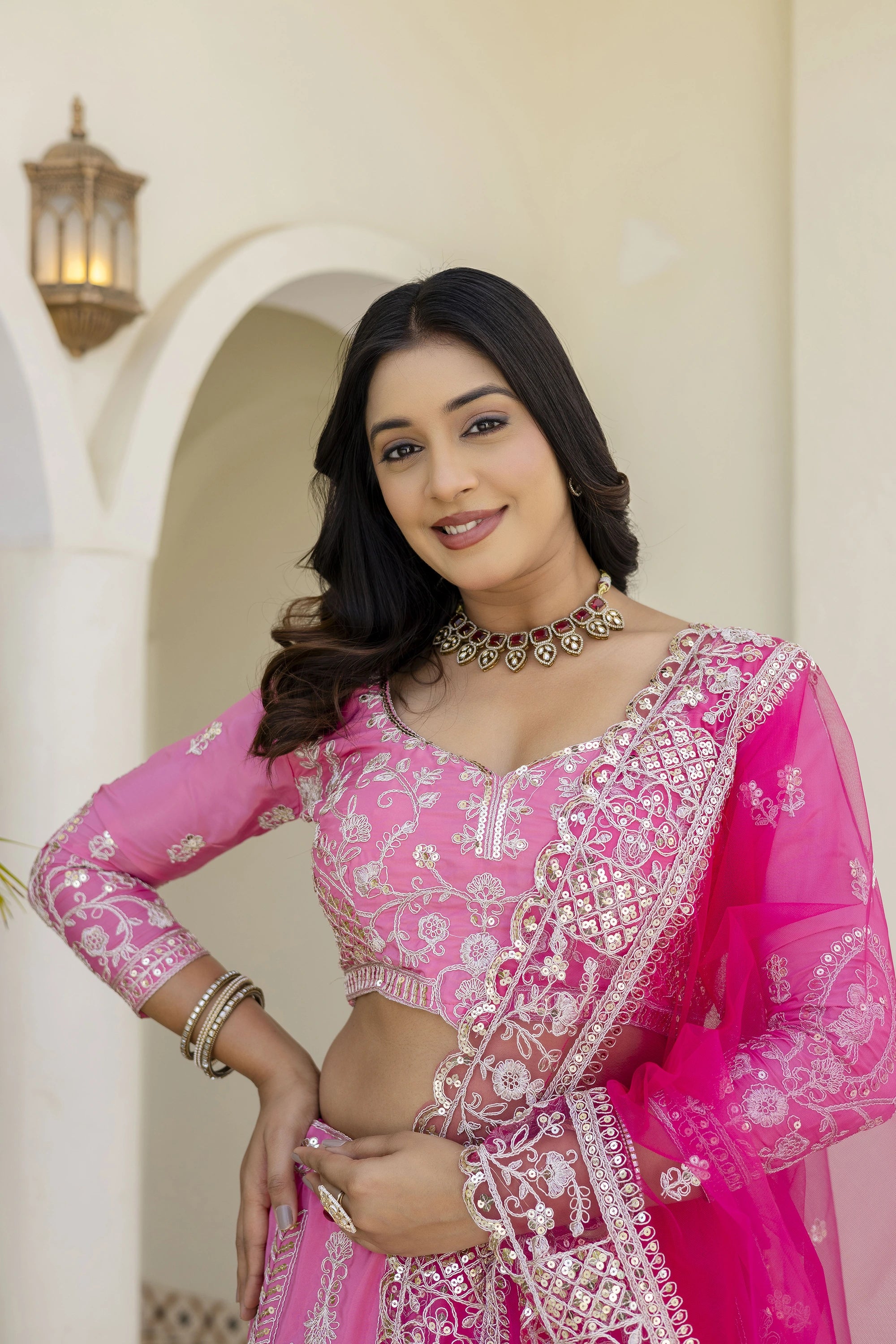Woman in a pink traditional outfit with intricate designs, standing against a light-colored wall.