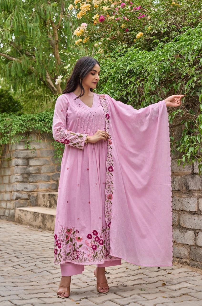 Woman in a pink traditional outfit with floral patterns standing outdoors near greenery.