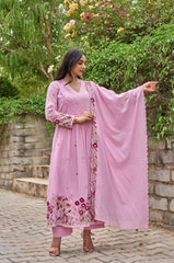 Woman in a pink traditional outfit with floral patterns standing outdoors near greenery.