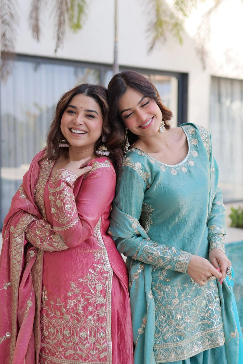 Two women in traditional pink and blue embroidered dresses standing together outdoors.