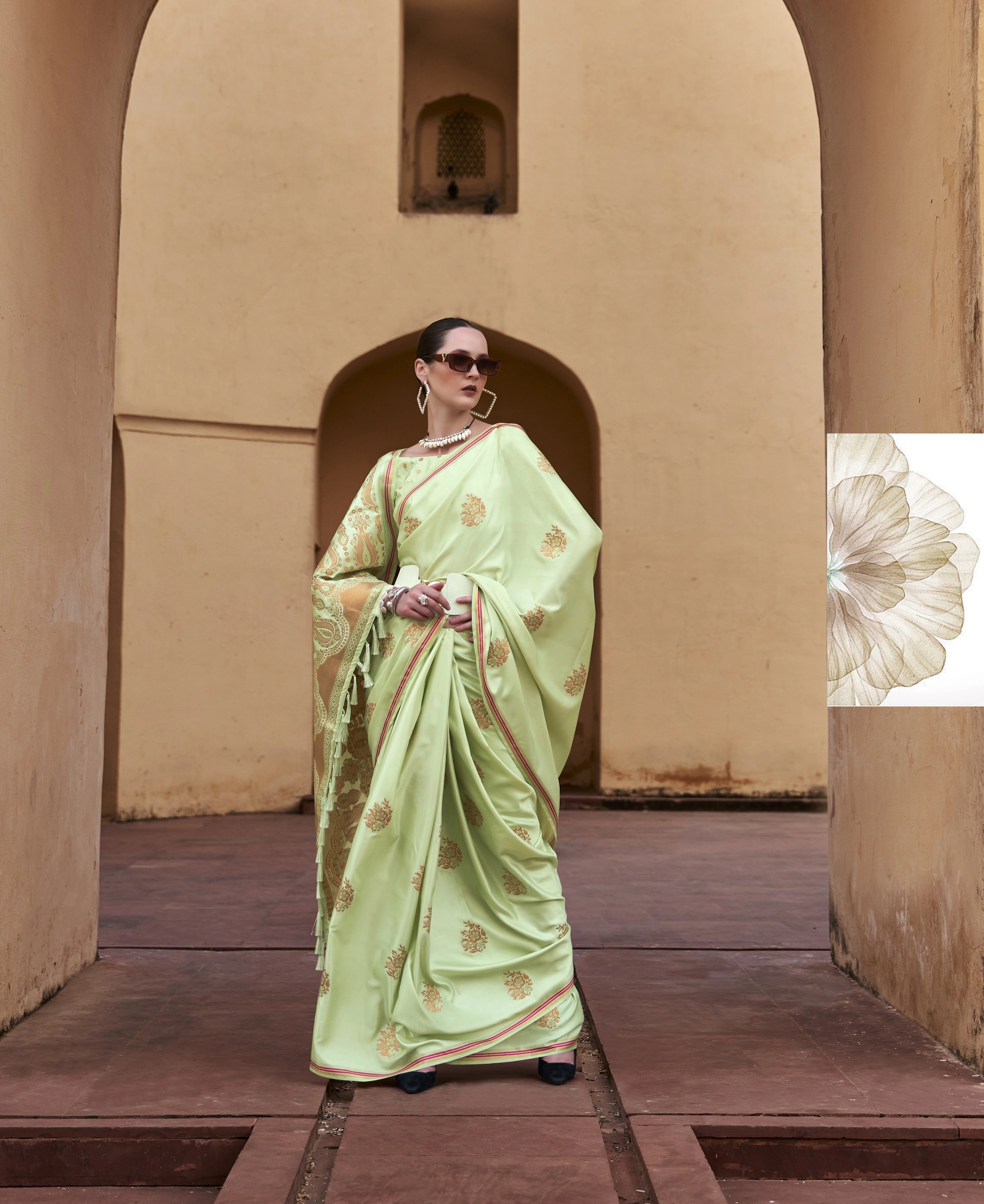 Woman in a light green saree standing in an archway with a beige wall and wooden floor.