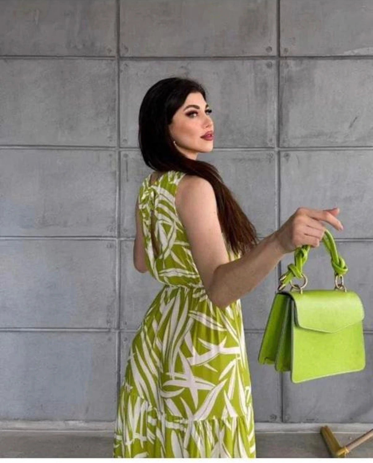 Woman in a green and white dress holding a lime green handbag against a gray tiled wall.