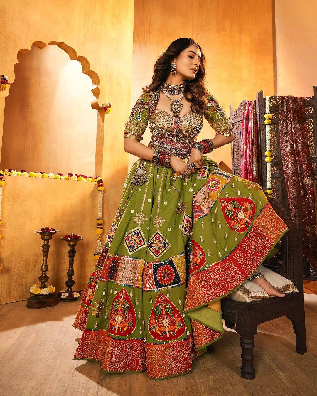 Woman in a traditional green and red outfit with intricate patterns in a decorated room.