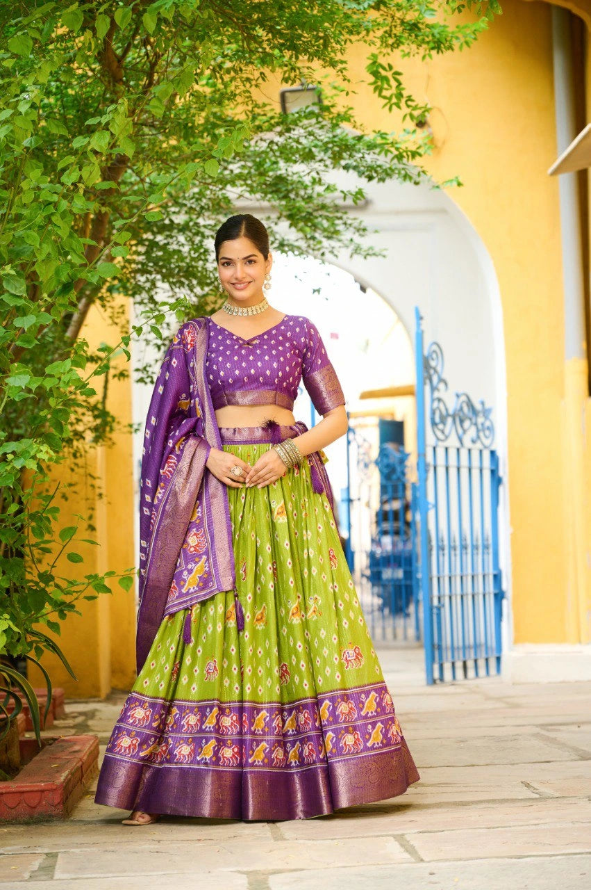 Woman in a green and purple traditional outfit standing outdoors with a yellow building and blue gate in the background.