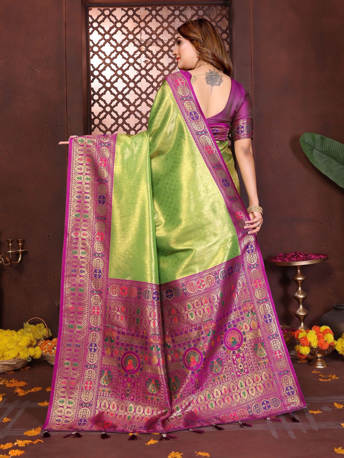 Woman wearing a green and purple saree with a decorative pattern, standing against a brown wall.